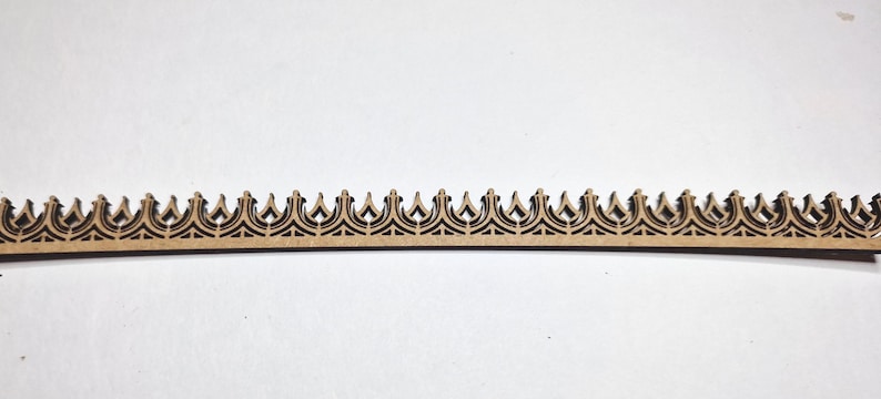 May include: A laser-cut wooden decorative trim piece with a repeating crown design. The trim is a light brown color and features intricate cut-out patterns. The piece is ideal for craft projects and home decor.