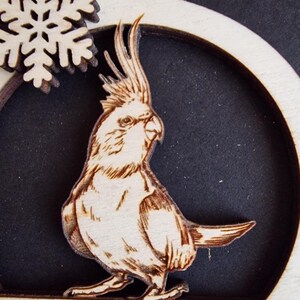 Puede incluir: Adorno navide&ntilde;o de madera con un grabado detallado de un cacat&uacute;a. El ave se encuentra sobre un fondo oscuro dentro de un marco circular, con un copo de nieve y texto en la parte superior. El adorno est&aacute; hecho de madera clara.