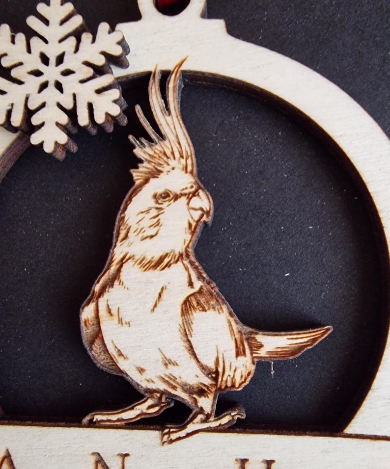 Puede incluir: Adorno navide&ntilde;o de madera con un grabado detallado de un cacat&uacute;a. El ave se encuentra sobre un fondo oscuro dentro de un marco circular, con un copo de nieve y texto en la parte superior. El adorno est&aacute; hecho de madera clara.