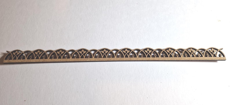 May include: A decorative wooden trim piece with an intricate, repeating arched design. The trim is a light brown color and features cut-out floral and curved patterns. The piece is long and narrow, suitable for craft projects or home decor.