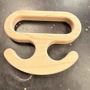 Puede incluir: Un asa de madera de color claro con un agarre ovalado y dos ganchos curvos debajo. El asa está hecha de madera sin terminar, mostrando la veta natural. El diseño sugiere que podría usarse para transportar o colgar objetos.