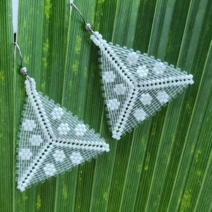 Pode incluir: Um par de brincos triangulares brancos com contas e um padrão geométrico. Os brincos são suspensos por ganchos prateados e são colocados sobre um fundo de folhas verdes. Os brincos são feitos de pequenas contas quadradas.