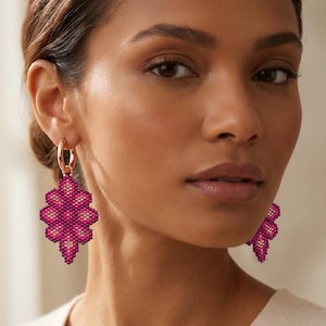 Può includere: Orecchini a cerchio in oro rosa con un pendente geometrico di perline nei toni del rosa e del crema. Gli orecchini hanno un design floreale e sono indossati da una persona.