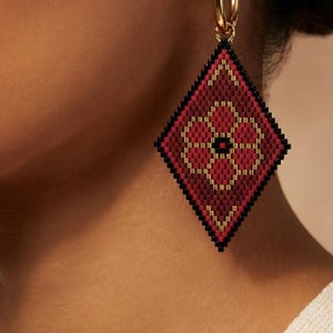 May include: Gold hoop earrings with a diamond-shaped pendant. The pendant is a mosaic of red, black, and gold beads, featuring a floral design in the center. The earrings are a striking accessory.
