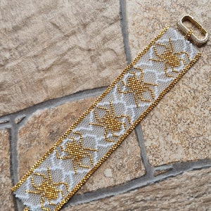 Op de afbeelding: Een witte en gouden kralen armband met een geometrisch patroon. De armband heeft een gouden sluiting.