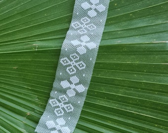 Peut inclure: Bracelet en perles blanches avec un motif floral, doté de fermoirs argentés. Le bracelet est posé sur une feuille de palmier verte.