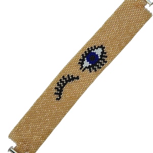 May include: A wide beaded bracelet featuring a gold-toned background with a central graphic design of an open eye and a closed eye with long eyelashes. The eye motif is rendered in shades of blue, white, and black beads, finished with silver-toned metal clasps.