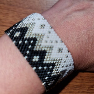 May include: A black and white beaded bracelet with a geometric pattern. The bracelet is worn on a person's wrist.