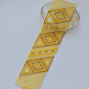 Può includere: Un braccialetto di perline bianche e gialle con un motivo geometrico. Il braccialetto ha una chiusura dorata.