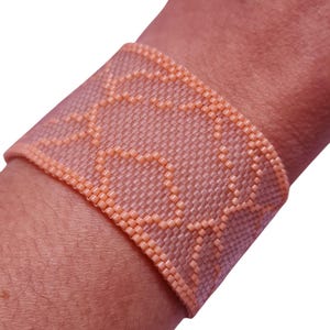Puede incluir: Un brazalete de cuentas de color melocotón con un patrón geométrico. El brazalete está hecho de pequeñas cuentas redondas que se tejen juntas para crear un diseño delicado e intrincado.