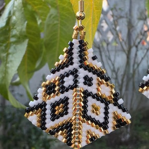 Puede incluir: Pendientes hechos a mano con cuentas y un patrón geométrico. Los pendientes tienen forma de pirámide con una combinación de cuentas blancas, negras y doradas. Cuelgan de ganchos dorados, mostrando un diseño único y artístico.