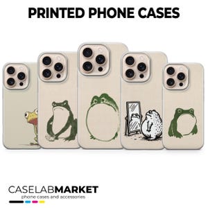 以下が含まれることがあります： さまざまなカエルのイラストが描かれた携帯電話ケースのコレクション。ケースはベージュ色で、縁は透明です。カエルのデザインは緑と黒です。「PRINTED PHONE CASES」の文字が上部にあります。