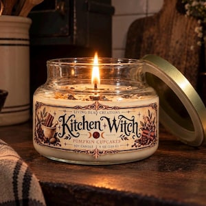 May include: A clear glass jar soy candle with a lit wick and a gold lid. The label features the text Living Dead Creations Kitchen Witch Pumpkin Cupcakes 8 oz (226 g) with illustrations of cinnamon sticks, a mortar and pestle, and moon phases.