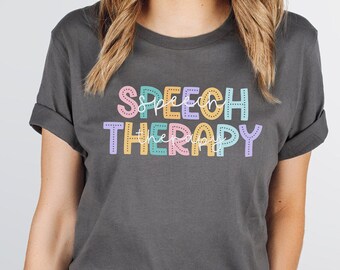 Puede incluir: Una camiseta gris oscuro con el texto "SPEECH THERAPY" en letras de colores y contorneadas. Las letras son en tonos de rosa, amarillo, azul y verde.