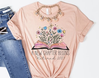 May include: A peach-coloured t-shirt with a graphic of an open book with flowers growing out of it. The text "A New Chapter Begins Retired 2023" is printed on the shirt.