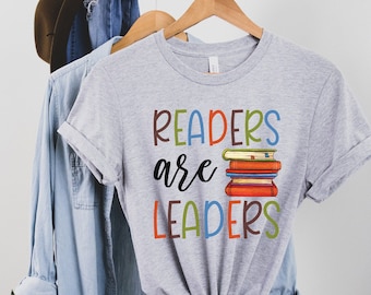 Puede incluir: Camiseta gris con el texto "Readers are Leaders" en letras de colores. Una pila de libros coloridos está al lado del texto.