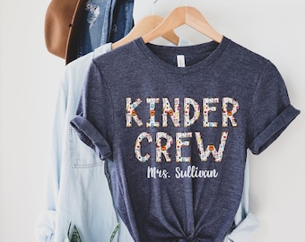 May include: A dark blue t-shirt with the text "Kinder Crew" in colorful letters with a school supply pattern. The text "Mrs. Sullivan" is printed below the main text.