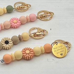 May include: Four colorful beaded keychains with gold metal clasps. Each keychain features a different color scheme and a flower charm. One keychain has a gold charm with the text "Never forget the difference you make!"