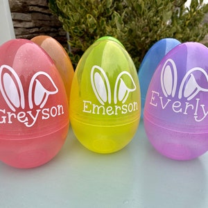 May include: A collection of colorful, glittery plastic Easter eggs. Each egg is a different color: pink, orange, yellow, blue, and purple. White bunny ears and a name are printed on each egg. The names are Greyson, Emerson, and Everly.