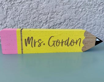 Puede incluir: Un bloque de madera con forma de lápiz que presenta un extremo de goma rosa, un cuerpo amarillo y una punta de madera natural con un extremo negro. El texto Mrs. Gordon está impreso en una fuente cursiva marrón oscuro sobre la sección amarilla.