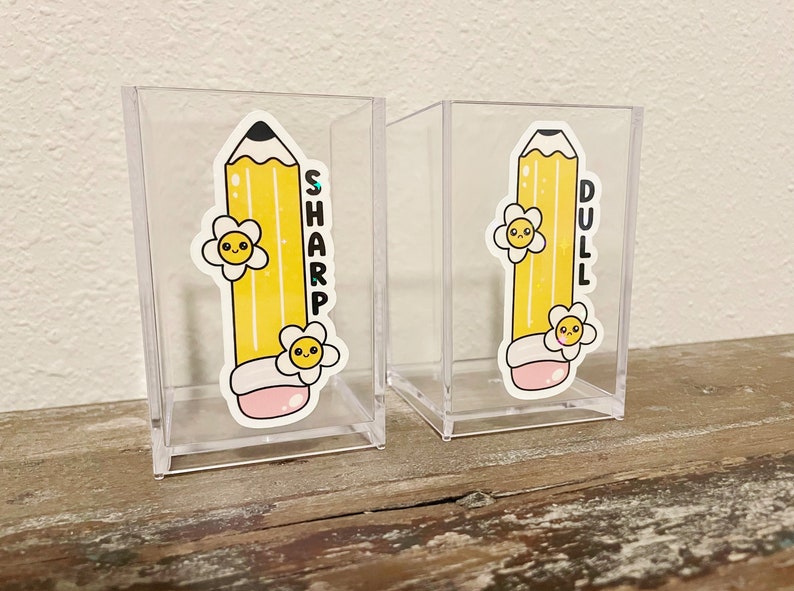 May include: Two clear acrylic pencil holders with yellow pencil stickers. One pencil holder has the word "SHARP" on it and the other has the word "DULL" on it.