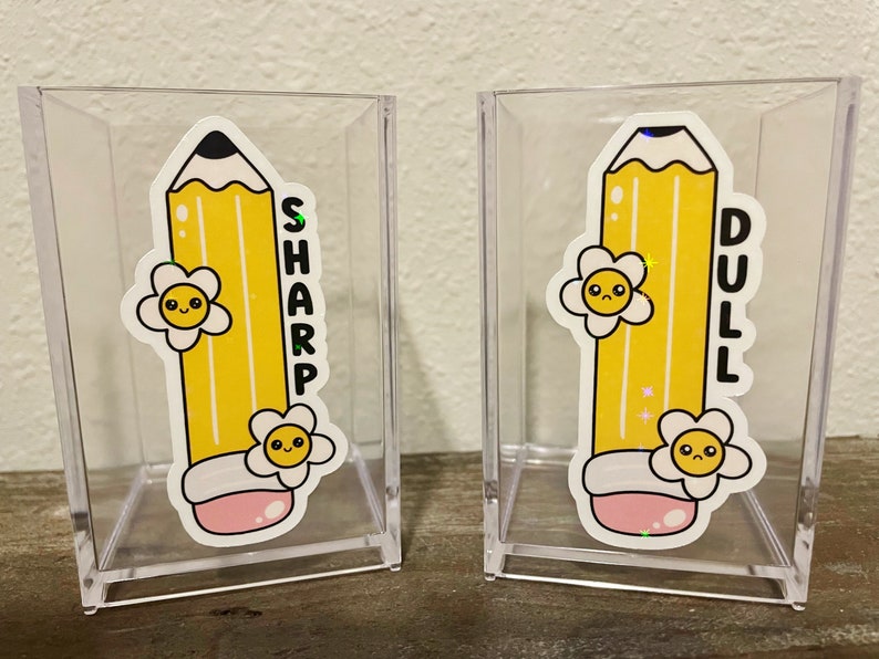 May include: Two clear acrylic pencil holders with yellow pencil stickers. The pencil on the left holder has a happy face and the word "SHARP" written vertically. The pencil on the right holder has a sad face and the word "DULL" written vertically.