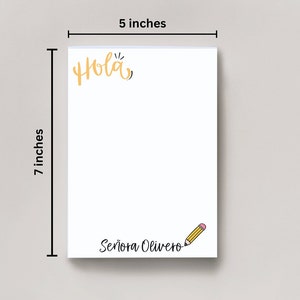 Op de afbeelding: Een wit notitieboekje met een gele "Hola" bovenaan en een geel potlood met een roze gum aan de onderkant. Het notitieboekje is 17,8 cm hoog en 12,7 cm breed. De tekst "Se&ntilde;ora Olivero" staat in zwart onderaan het notitieboekje.