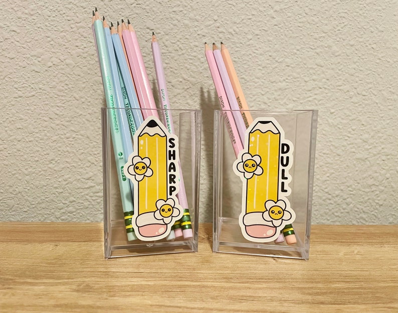 May include: Two clear plastic pencil holders with colorful pencils inside. One holder has a sticker of a cartoon pencil with the word "SHARP" and the other has a sticker of a cartoon pencil with the word "DULL".