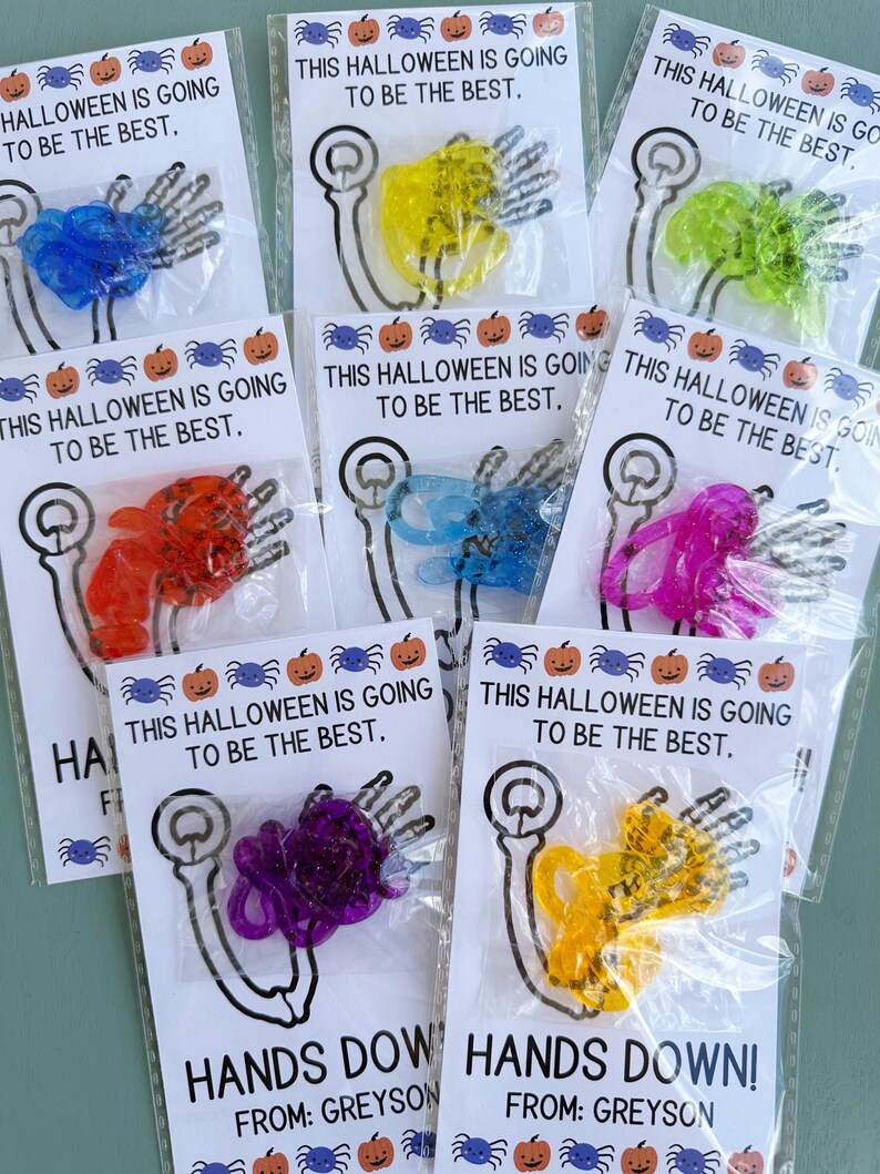 May include: Six small bags with a white card inside each bag. The card says "This Halloween is going to be the best." and has a drawing of a skeleton arm. Each bag contains a different color of plastic spider rings. The bags are labeled "Hands Down!" and "From: Greyson".