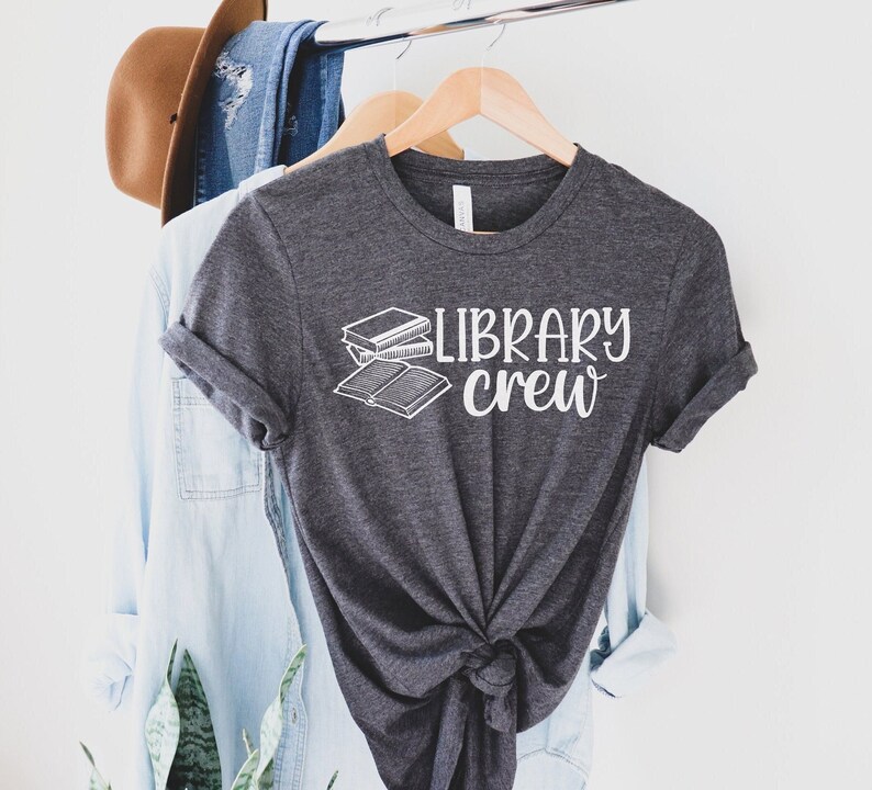 May include: A charcoal grey t-shirt featuring the white text "LIBRARY CREW" and a book illustration. The tee is hanging on a wooden hanger, with other clothes and a brown hat visible in the background.