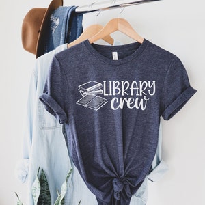 May include: A navy blue t-shirt with the words "LIBRARY crew" in white script and a graphic of two books. The shirt is hanging on a wooden hanger with other clothing items in the background.