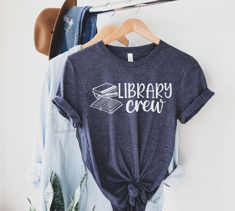 May include: A navy blue t-shirt with the words "LIBRARY crew" in white script and a graphic of two books. The shirt is hanging on a wooden hanger with other clothing items in the background.