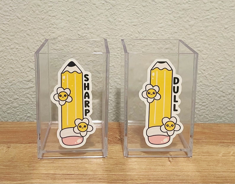 May include: Two clear plastic pencil holders with yellow pencil stickers. The pencil on the left holder has a happy face and the text "SHARP". The pencil on the right holder has a sad face and the text "DULL".