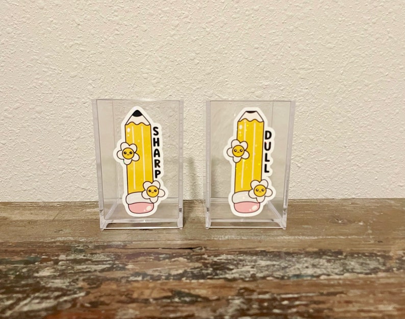 May include: Two clear acrylic pencil holders with yellow pencil stickers. The pencil on the left holder has the word "SHARP" written vertically on it. The pencil on the right holder has the word "DULL" written vertically on it.