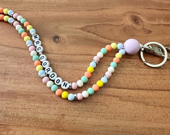 May include: A colorful beaded lanyard with the name "MRS GORDON" spelled out in black letters on white beads. The lanyard has a purple bead at the end and a silver key ring.