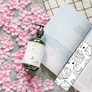 May include: A green glass bottle of body wash with a white label that says "KIM THOA" and "DƯỠNG ẨM DA NHẠY CẢM HOA & VO BUC". The bottle is sitting on a surface covered in pink flower petals.