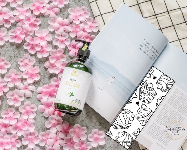 May include: A green glass bottle of body wash with a white label that says "KIM THOA" and "DƯỠNG ẨM DA NHẠY CẢM HOA & VO BUC". The bottle is sitting on a surface covered in pink flower petals.