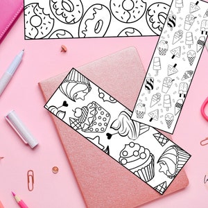 May include: Three black and white printable bookmark designs featuring cupcakes, donuts, and ice cream cones. The bookmarks are on a pink surface with a pink notebook, pencils, and paper clips.