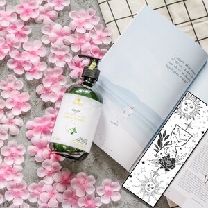May include: A green glass bottle with a white label that says "Dầu Gội" and "HOA & VO NUC" in black text. The bottle is sitting on a surface covered in pink flower petals. A black and white bookmark with a sun, moon, and stars design is visible in the background.