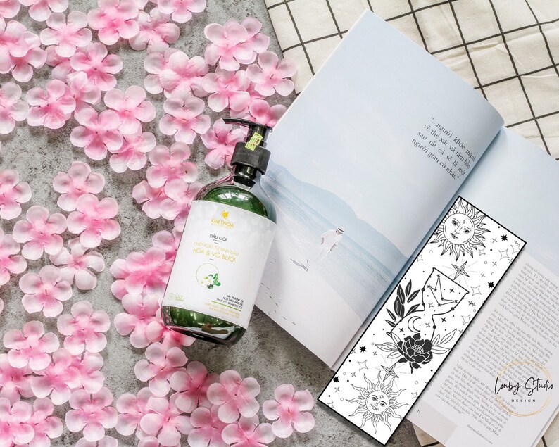 May include: A green glass bottle with a white label that says "Dầu Gội" and "HOA & VO NUC" in black text. The bottle is sitting on a surface covered in pink flower petals. A black and white bookmark with a sun, moon, and stars design is visible in the background.