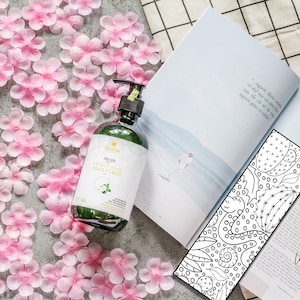 Puede incluir: Una botella de gel de ducha de vidrio verde con una etiqueta blanca que dice "Dầu Gội HOA & VO BUỔI". La botella est&aacute; sobre una superficie blanca con p&eacute;talos de flores rosas esparcidos alrededor.
