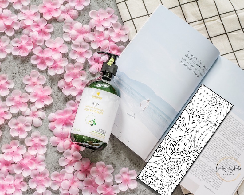 Puede incluir: Una botella de gel de ducha de vidrio verde con una etiqueta blanca que dice "Dầu Gội HOA & VO BUỔI". La botella est&aacute; sobre una superficie blanca con p&eacute;talos de flores rosas esparcidos alrededor.