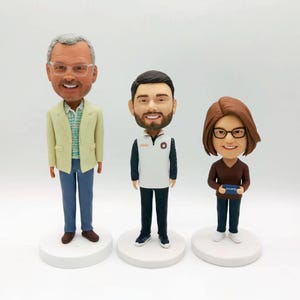 Könnte beinhalten: Drei personalisierte Wackelköpfe, einer mit einer hellgrünen Jacke, einer mit einem weißen Hemd und einer schwarzen Hose und einer mit einem braunen Hemd und einer schwarzen Hose. Die Wackelköpfe stehen auf weißen Sockeln.