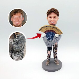 May include: A custom bobblehead figurine of a person wearing camouflage pants and a grey shirt holding a turkey fan. The bobblehead has a realistic face and is a great gift for hunters.