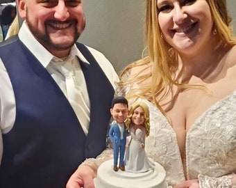 May include: A white wedding cake with a fondant topper of a bride and groom. The bride is wearing a white dress and the groom is wearing a blue suit. The cake is on a white plate and is being held by a bride and groom.