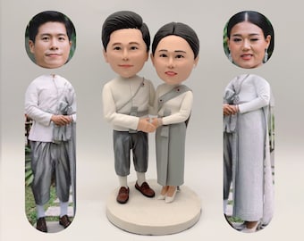 May include: A custom bobblehead couple wearing traditional Thai clothing. The male bobblehead is wearing a white shirt with a gray sash and gray pants. The female bobblehead is wearing a white shirt with a gray sash and a gray skirt. The bobbleheads are standing on a white base.