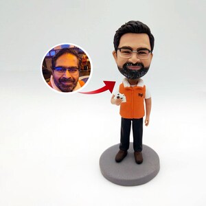 Könnte beinhalten: Eine personalisierte Bobblehead-Figur einer Person, die ein schwarzes Hemd, eine orange Weste und eine schwarze Hose trägt. Die Figur hält ein weißes Objekt in ihrer rechten Hand. Die Figur steht auf einem grauen Sockel. Das Gesicht der Person ist ein Foto eines Mannes mit Bart und Brille.