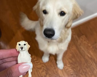 May include: A small, white, ceramic figurine of a dog sitting on a person's hand. The figurine is a replica of a larger, white dog standing in the background.