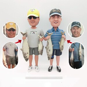 May include: Two custom bobbleheads of people holding fish. The bobbleheads are wearing casual clothing and hats. The bobbleheads are standing on a white base.