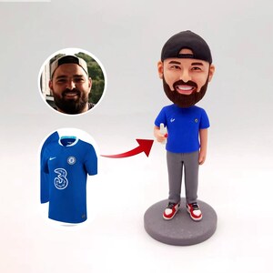 Könnte beinhalten: Eine personalisierte Bobblehead-Figur einer Person, die ein blaues Trikot mit einer weißen Nummer 3 auf der Vorderseite trägt. Die Figur hält ein kleines weißes Objekt in ihrer rechten Hand. Die Figur steht auf einem grauen Sockel.